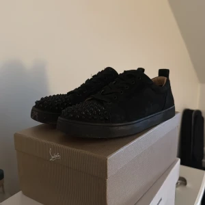 Svarta Christian Louboutin skor - Säljer ett par svarta sneakers från Christian Louboutin med nitar och klassisk röd sula. Skorna har snörning och är tillverkade i mocka med en platt sula. Säljer då jag knappt använder längre. En nit saknas på ena skon samt att de är synligt använda därav låga priset