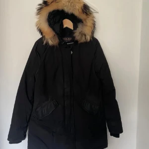 Svart parkas från Woolrich med päls - Parkas från Woolrich i svart med stor huva och fluffig päls i beige och bruna toner. Jackan har två snedställda fickor framtill och stängs med dragkedja och knappslå. Perfekt för kalla dagar med sin långa, varma design och klassiska look.