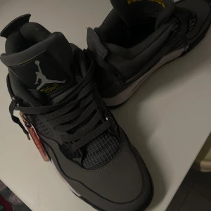 Nike Air Jordan 4 Retro Grå - Säljer ett par Nike Air Jordan 4 Retro sneakers i mörkgrått med svarta och vita detaljer. Skorna har klassisk snörning, meshpaneler och Jumpman-logga i silver på hälen. Ovandelen är i skinn och syntet, med platt sula och rund tå. Tag medföljer.