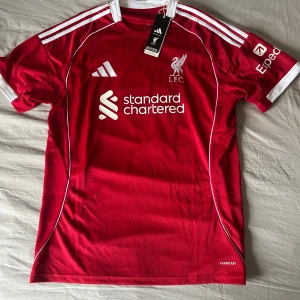 Liverpool röd fotbollströja Adidas L - Liverpool FC matchtröja från Adidas i rött med vita detaljer och tryck. Kortärmad modell med klassiska tre ränder på axlarna, klubbmärke och sponsortryck fram, samt ISAK och nummer 9 på ryggen. Tillverkad i lätt, ventilerande polyester med Aeroready-teknologi.