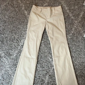 Beiga bootcut byxor med fickor - Snygga beiga byxor med bootcut-snitt och bred linning med knappar. De har fickor bak med lock och dekorativa detaljer framtill. 