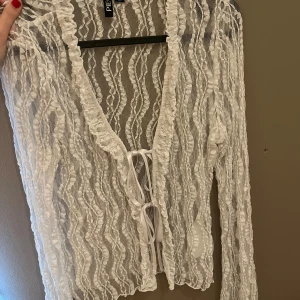 Vit bolerotopp med snörning - Säljer en vit bolerotopp från PIECES med transparent mesh och vågiga strukturer. Toppen har lång ärm, v-ringning och snörning framtill. Perfekt för att lägga till en extra touch till din outfit. Supersnygg att styla över linne eller klänning.