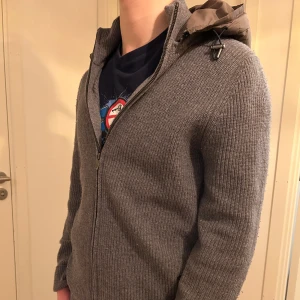 Grå Massimo Dutti jacka - Mysig grå stickad Massimo Dutti jacka med quiltad huva och dragkedja framtill. Jackan har en ribbad struktur och lång ärm, perfekt för kyligare dagar. Enkel och stilren design som passar till det mesta. Säljs inte längre så därför priset. 