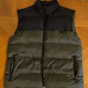 Svart/grön hugo boss väst - Snygg dunväst från Hugo boss i svart och olivgrönt med hög krage och två dragkedjeförsedda fickor. Helt ny/oanvänd är den. Västen har quiltad design och ett Hugo-märke på bröstet. Insidan är fodrad med Hugo-loggor och har en röd etikett. Perfekt för lager-på-lager under kyliga dagar. 