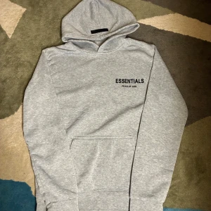 Grå Essentials Fear of God hoodie - Säljer en grå hoodie från Essentials Fear of God med svart logga på bröstet och ryggen. Tröjan har huva och en stor magficka framtill. Perfekt för dig som gillar streetwear och enkel stil.