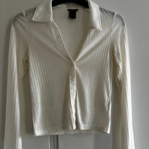 Vit ribbad kofta från Lindex XS - Säljer en stilren vit ribbad kofta från Lindex i storlek XS. Koftan har lång ärm, klassisk krage och knäppning framtill med fyra knappar. Perfekt att slänga över ett linne eller t-shirt för en clean look.