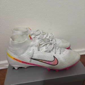 Nike fotbollsskor gjorde av Marcus rashford (Ag) - Säljer ett par Nike fotbollsskor i vitt med snygga rosa och gula detaljer samt texten 'When you believe in yourself' på sidan. Skorna har snörning, högre strumpliknande skaft och platt sula med dobbar i rosa och orange. Perfekta för dig som vill sticka ut på planen. Använda 2 gånger på träning så inge fel på de alls.