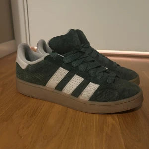 Adidas campus 00s Japanese Rock Garden Green Oxide - Säljer ett par Adidas campus 00s i mörkgrön mocka med klassiska vita ränder på sidorna och grå insida. Skorna har breda gröna snören och en brun sula. Perfekta för dig som gillar retrostil och vill sticka ut lite extra.