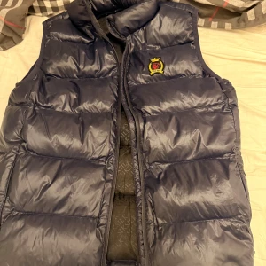 Mörkblå dunväst från SikSilk - Säljer en mörkblå dunväst från SikSilk med broderad logga på bröstet. Västen har quiltad design, hög krage och dragkedja framtill. Insidan är mönstrad med SikSilk-tryck. Perfekt för lager-på-lager under kyliga dagar.