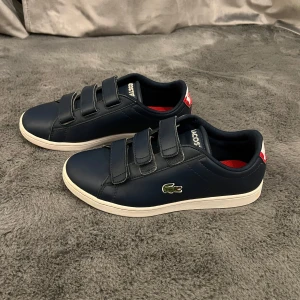 Mörkblå sneakers från Lacoste - Snygga mörkblå sneakers från Lacoste med tre kardborreband och vit platt sula. Skorna har klassisk rund tå och är tillverkade i skinn med röd detalj och krokodillogo på sidan. Insidan är röd och hälen har en vit och röd Lacoste-logga. 