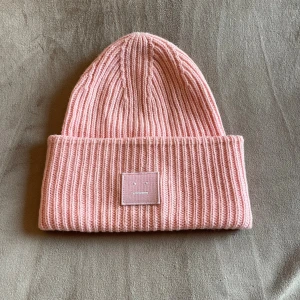 Rosa ribbad mössa Acne Studios - Snygg rosa mössa från Acne Studios med ribbad stickning och uppvikt kant. Perfekt accessoar för att lyfta din look under kyliga dagar. Något synliga smink fläckar i mössan men går att tvätta bort!