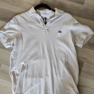 Vit pikétröja från Lacoste - Snygg vit pikétröja från Lacoste med klassisk krage och knappar framtill. Tröjan har korta ärmar och den ikoniska krokodilloggan på bröstet. Perfekt för dig som gillar stilrena och sportiga plagg.
