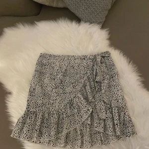 Prickig omlottkjol med volang - Säljer en kort omlottkjol med svartprickigt mönster på vit botten. Kjolen har volangkant nedtill och knyt i sidan för extra detalj. Perfekt för dig som gillar trendiga och lekfulla plagg med lite extra rörelse. Materialet känns lätt och luftigt.