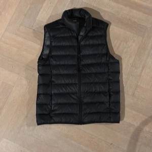 Svart dunväst från Uniqlo XS - Svart dunväst från Uniqlo Ultra Light Down i storlek XS. Västen har en stilren, quiltad design med ståkrage och dragkedja framtill. Tillverkad i lätt och vattenavvisande syntetmaterial, perfekt för lager-på-lager under kyliga dagar.