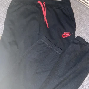 Svarta mjukisbyxor från Nike med röd logga - Svarta mjukisbyxor från Nike med röd broderad logga på benet och matchande röd snörning i midjan. Byxorna har elastisk midja och är gjorda i mjukt bomullsmaterial, perfekta för chill eller träning. Passar mig som bär st xs 💗