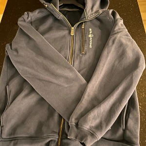 Mörkblå hoodiejacka från Sail Racing - Mörkblå hoodiejacka från Sail Racing med guldfärgade dragkedjor och broderad logga på bröstet och huvan. Jackan har lång ärm, huva och flera fickor. Materialet är mjukt och känns som bomull, perfekt för chill dagar.