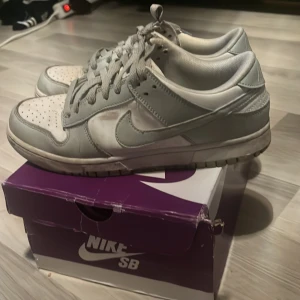 Nike Dunk Low i grått och vitt - Nike Dunk Low sneakers i en clean grå och vit färgkombination. Skorna har klassisk snörning, perforerad tåbox och ikonisk Swoosh på sidorna. Tillverkade i skinn med platt sula och rund tå. Perfekta för dig som gillar streetwear och vill ha en tidlös look.