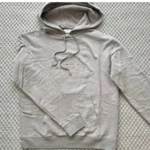 Grå hoodie från Saint Laurent - Snygg grå hoodie från Saint Laurent med huva och snörning. Tryck med små stjärnor och logga på bröstet. Kan uppfattas som något beige i verkligheten. Mjuk bomullskänsla och klassisk känguruficka framtill. Priset ej hugget i sten!