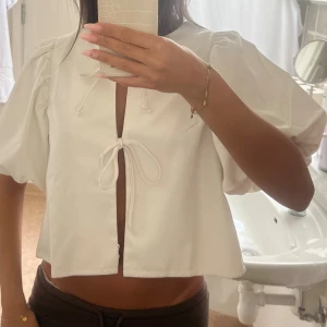 Vit blus med puffärm och knyt - Säljer en vit blus med korta puffärmar och öppen front som knyts med två band. Blusen är croppad och har en luftig passform, perfekt för varmare dagar. Materialet känns mjukt och lätt, och designen ger en trendig och söt look.