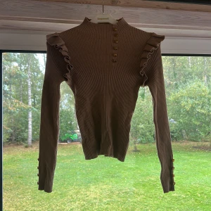 Beige ribbad topp med volang och knappar - Supersnygg beige ribbad topp med lång ärm, volangdetaljer på axlarna och guldknappar både framtill och vid ärmsluten. Hög hals och figurnära passform gör den extra trendig. Perfekt att styla med jeans eller kjol för en cool look.