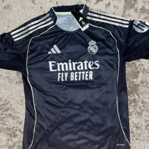 Real Madrid Mbappé tröja Adidas L - Säljer en mörkblå Real Madrid fotbollströja från Adidas i storlek L med Mbappé och nummer 10 på ryggen. Tröjan har korta ärmar, silverfärgade ränder på axlarna och klubbmärke på bröstet. Tillverkad i lätt och ventilerande material, perfekt för match eller träning.