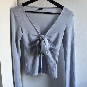 Ljusgrå långärmad topp Gina Tricot - Supersnygg ljusgrå långärmad topp från Gina Tricot med v-ringning och knyt framtill. Tunn och mjuk i materialet, perfekt att styla med jeans eller kjol. Enkel men ändå trendig design som ger en clean look.
