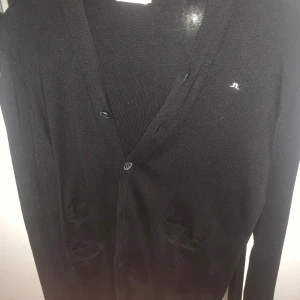 Svart cardigan från J.Lindeberg - Svart kofta från J.Lindeberg med diskret logga på bröstet. Koftan har v-ringning, knappar framtill och två fickor. Långärmad och tillverkad i mjukt material som känns skönt mot huden. Perfekt för lager-på-lager och chill stil. Jag är 185 cm och den är lite för kort för mig👍🏻