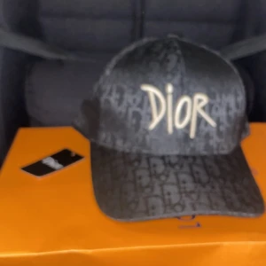 Dior keps  - Snygg svart keps från Dior med ikoniskt logomönster över hela kepsen och broderad Dior-text framtill i beige. Klassisk böjd skärm och justerbar passform. Perfekt accessoar för att lyfta din streetwear-look.