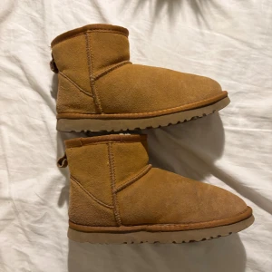 UGG Classic Mini bruna boots i mocka - UGG Classic Mini boots i ljusbrun mocka med rund tå och platt sula. Insidan är fodrad med mjuk, vit päls för extra värme. Perfekta för kalla dagar och har den klassiska UGG-loggan under sulan.