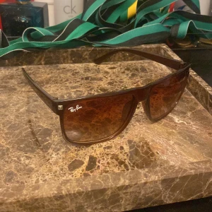 Bruna Ray-Ban solglasögon - Snygga solglasögon från Ray-Ban med brun fyrkantig plastbåge och tonade bruna glas. Klassisk logga på vänster sida och cool, bred design som ger en modern vibe. Perfekta för att höja din stil på soliga dagar.