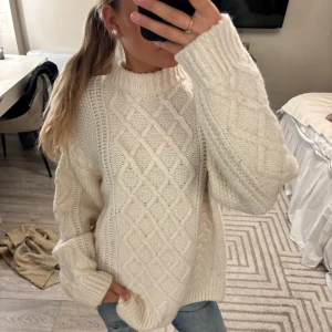 Vit stickad tröja med flätmönster - Supermysig vit stickad tröja med grovt flätmönster och ribbad hög hals. Tröjan har en oversized passform och långa ärmar, perfekt för kyliga dagar. Snygg att styla med jeans eller leggings för en chill look.