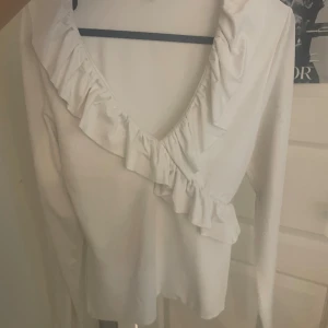 Vit blus med volang och v-ringning - Säljer en vit blus med djup v-ringning och snygg volangdetalj längs ringningen. Blusen har långa ärmar och är perfekt för dig som gillar en feminin och stilren look. Passar till både jeans och kjol för en trendig outfit.