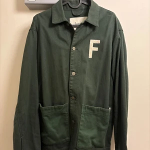 Club Overshirt Dark Green från Forét  - Mörkgrön overshirt från Forét med stora fickor framtill och broderad text på ryggen. Framsidan har en vit broderad bokstav F och tryckknappar. 