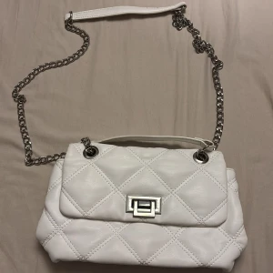 Vit quiltad axelväska med kedja - Snygg vit axelväska med quiltat mönster och silverfärgad kedjerem. Väskan har ett klassiskt lock med vridspänne i metall och mjukt material. Perfekt för dig som vill ha en stilren och trendig accessoar.