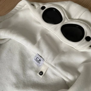 Vit hoodie från C.P. Company - Säljer en vit hoodie från C.P. Company med ikoniska goggles på luvan. Helt nytt skick, har andvänt 1 gång. Nypris: 3399kr, mitt pris kan självklart alltid diskuteras!🙌💯⚜️