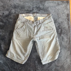 Randiga blåvita shorts i bomull, strl 32 - Snygga shorts med smala blå och vita ränder, klassisk design och normal passform. Tillverkade i mjuk bomull med fickor fram och bak, samt knappstängning bak. Perfekta för dig som gillar chill och stilren look.