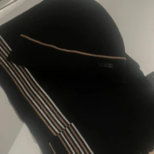 BOSS mössa och halsduk  - Svart mössa och halsduk från BOSS med en beige och vit rand längs uppviket. Stickad i mjukt material som känns skönt mot huden. Snygg metallbricka med BOSS-logga framtill. Perfekt för dig som gillar stilrena accessoarer med diskreta detaljer.