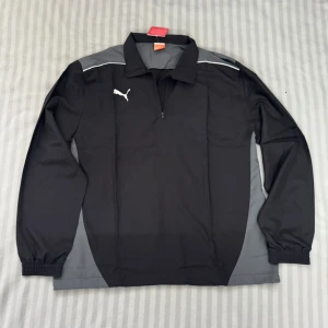 Svart sporttröja från Puma XXL - Svart och grå långärmad sporttröja från Puma med halv dragkedja och vit logga på bröstet. Tröjan har elastiska muddar och är tillverkad i 100% polyester, perfekt för träning eller fotboll. Snygg kontrast mellan svart och grått med diskreta vita detaljer. Aldrig använts