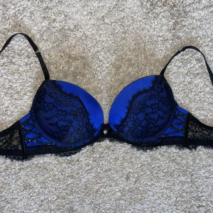 Blå och svart bh med strass och spets - Snygg bh i klarblå satin med svarta spetsdetaljer och tunna axelband. Den har dekorativa svarta band, små strassdetaljer och en söt rosett med glittrande sten framtill.  75A💕