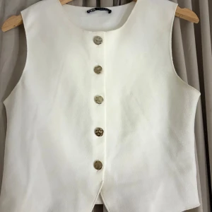 Vit väst från Zara med snygga knappar - Stilren vit väst från Zara i storlek M. Västen har rund halsringning och fem unika, hamrade knappar framtill. Materialet är mjukt och har en lätt struktur som ger en exklusiv känsla. Perfekt för att lyfta din outfit med en clean och modern look.