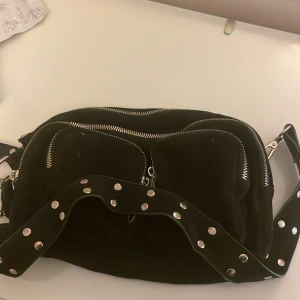 Svart axelväska med nitar från Gina Tricot - svart axelväska från Gina Tricot med bred axelrem dekorerad med silvriga nitar. Väskan har flera fack med dragkedjor och är tillverkad i ett mjukt syntetmaterial.