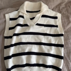 Randig stickad väst med krage - Snygg stickad väst i vitt med svarta horisontella ränder och en bred, ribbad krage. Västen är ärmlös och har en v-ringad halsringning. Perfekt att styla över en skjorta eller t-shirt för en trendig look.