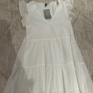 Vit sommarklänning från H&M Divided - Superfin vit klänning från H&M Divided med volangärmar och v-ringning. Tillverkad i lätt och luftig polyester, perfekt för varma dagar. Klänningen har en flowy passform och flera sömmar som ger en snygg siluett.