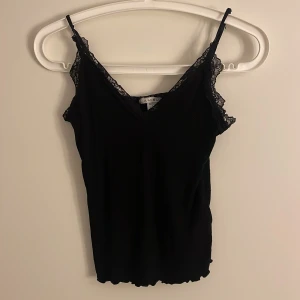 Svart linnetopp med spets  - Svart linnetopp med tunna axelband och fina spetsdetaljer längs urringning och axlar. Toppen har en lätt v-ringning och vågig nederkant, perfekt att styla till jeans eller kjol. Mjuk och följsam känsla.