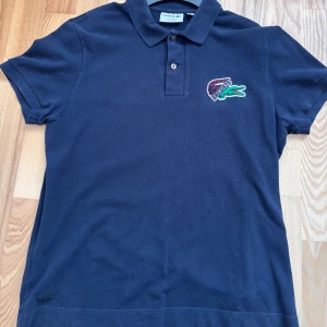 Mörkblå Lacoste pikétröja i bomull - Snygg mörkblå pikétröja från Lacoste med klassisk krage och två knappar framtill. Tröjan har korta ärmar och är tillverkad i bomull. På bröstet finns den ikoniska krokodilloggan i färg och en ton-i-ton krokodil nertill. Regular fit.