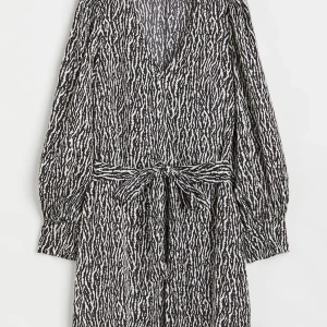 Svartvit omlottklänning med zebra-mönster - Snygg kort omlottklänning i svartvitt zebramönster med långa ärmar och knytband i midjan. Djup v-ringning och lätt, flowigt material som ger en cool och trendig look. Perfekt för dig som vill sticka ut med ett djurmönster.