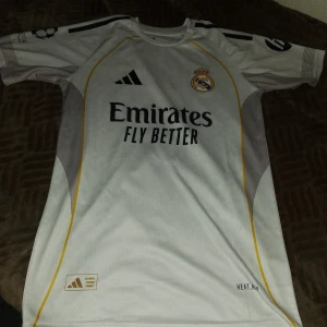 Real Madrid Vini Jr. 7 matchtröja Adidas - Säljer en officiell Real Madrid fotbollströja från Adidas med Vini Jr. och nummer 7 på ryggen. Tröjan är vit med svarta detaljer, gula linjer och klubbmärke på bröstet. Champions League-märke på ärmen och tillverkad i lätt funktionsmaterial.