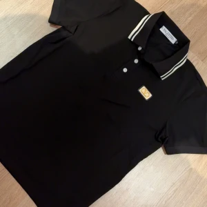 Svart pikétröja från Valentino - Svart pikétröja från Valentino med klassisk krage och vita ränder. Tröjan har korta ärmar, tre knappar framtill och en snygg guldfärgad logotyp på bröstet. Tillverkad i mjuk bomull för en skön känsla och stilren look.