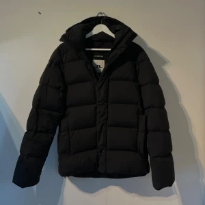 Svart pufferjacka från J.Lindeberg - Säljer en svart pufferjacka från J.Lindeberg med huva och dragkedja. Jackan har en clean, stilren look med diskret logga på ärmen. Perfekt för kalla dagar och enkel att matcha med allt. Andvänt skick men inte trasig eller några hål.