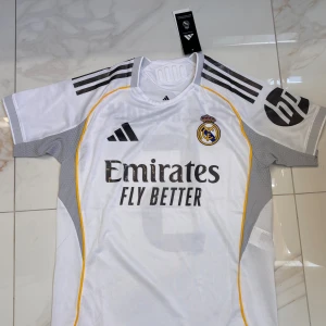 Real Madrid Bellingham - Snygg vit Real Madrid fotbollströja från Adidas med Bellingham och nummer 5 på ryggen. Tröjan har svarta och grå detaljer, gula linjer längs sidorna och klubbmärke på bröstet. Tillverkad i lätt och ventilerande polyester, perfekt för match eller träning.